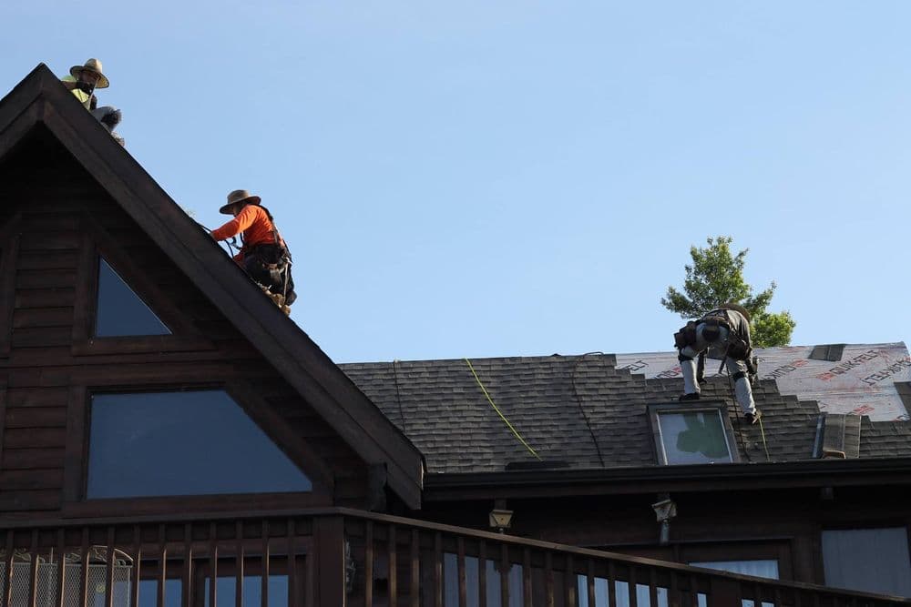 Roofers installing shingles on a house, one using a robotic assistive device for safety.