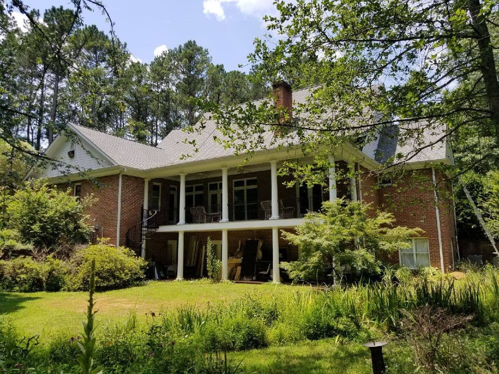 Two-story brick house with a porch, surrounded by greenery and trees. Sunny day, blue sky.