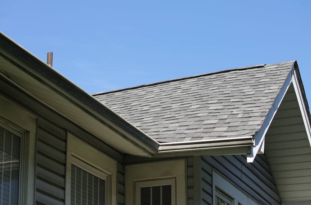 Close-up of a gray shingle roof meeting a house's exterior under a clear blue sky.