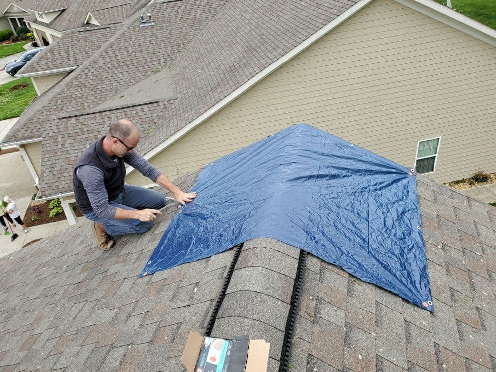 Roof repair worker securing a blue tarp on a sloped roof to prevent water damage.