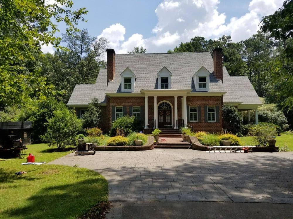 Large brick house with a distinctive roof, surrounded by greenery and a paved driveway.