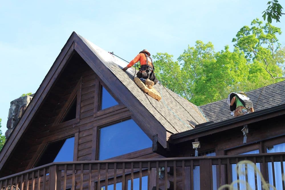 Roofers working on a wooden cabin's roof under clear blue skies. Trees in the background.