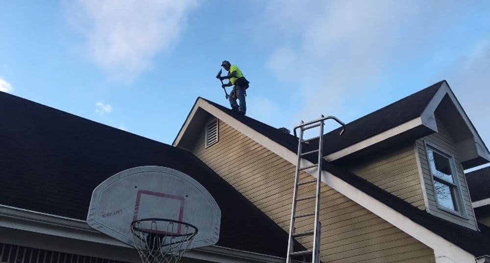 Worker repairing a roof with a ladder and basketball hoop nearby under a blue sky.