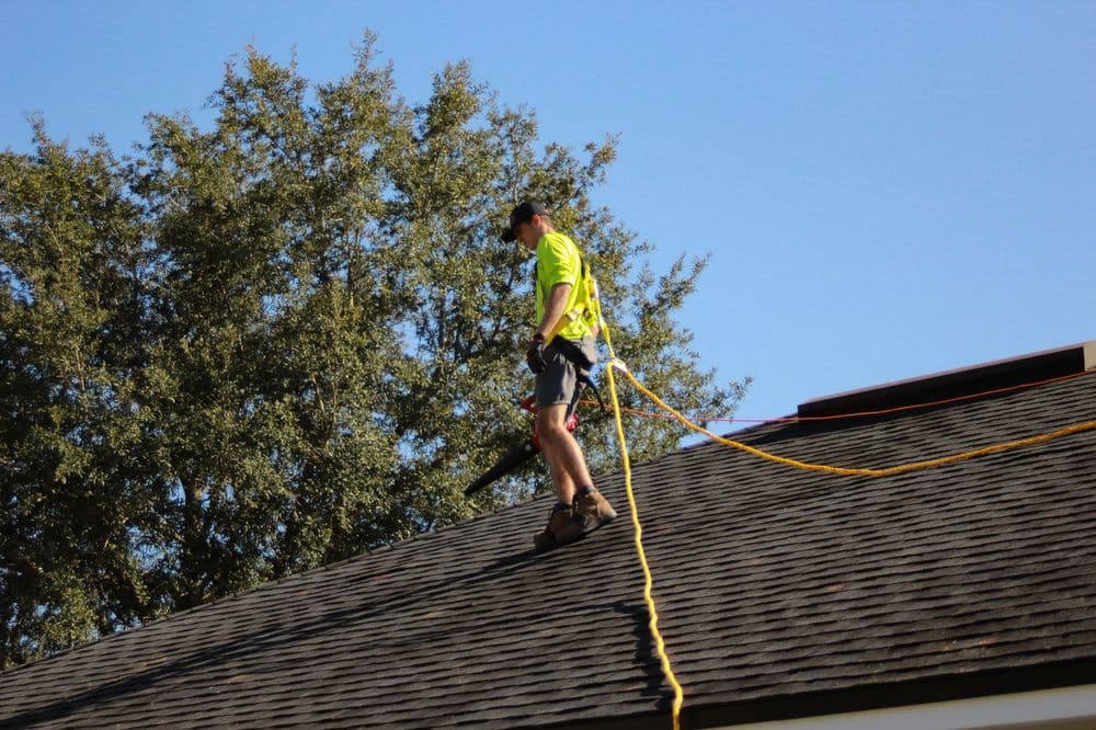 Worker in neon shirt using a leaf blower while standing on a sloped roof. Safety harness visible.