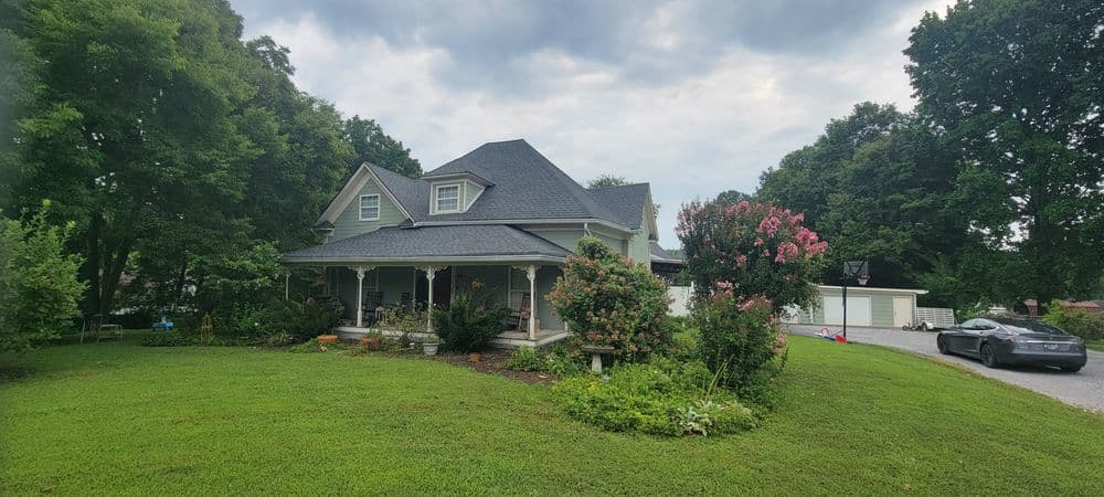 Victorian-style house with lush garden and trees, featuring a wraparound porch and driveway.