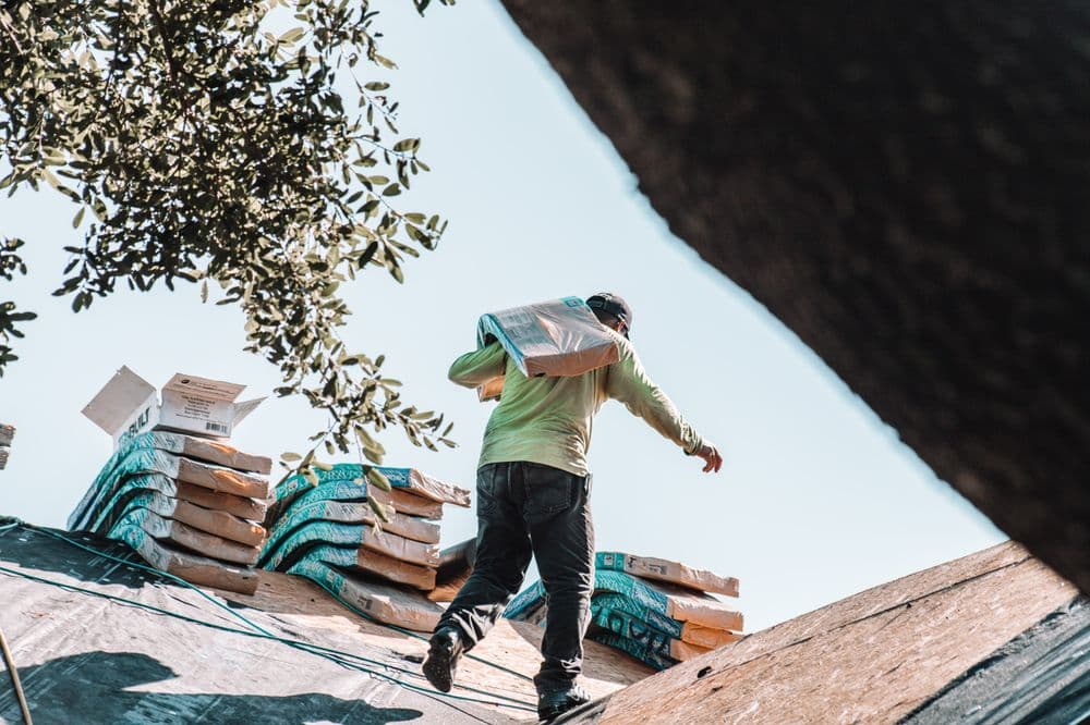 Worker carrying bags on a roof, surrounded by stacked construction materials and trees.