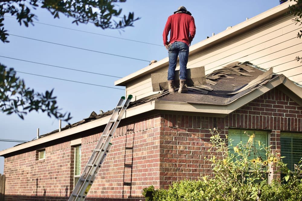 Roofer standing on a house roof, preparing to install shingles with a ladder nearby.